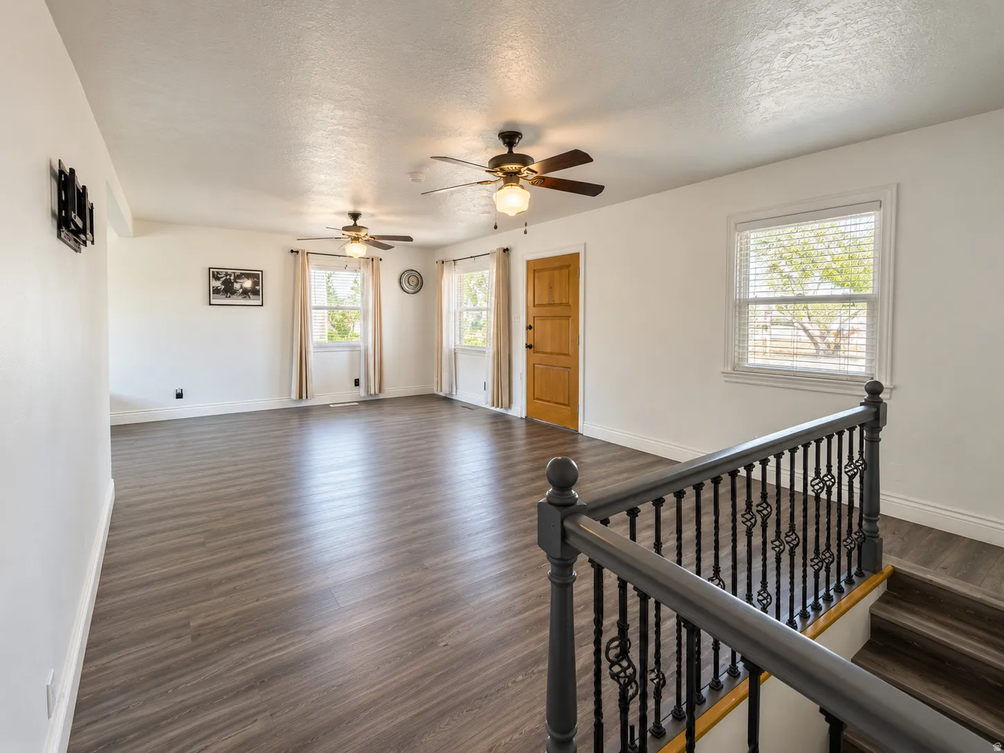 Spacious living area featuring wood-finish flooring, multiple windows, and two ceiling fans with integrated lighting