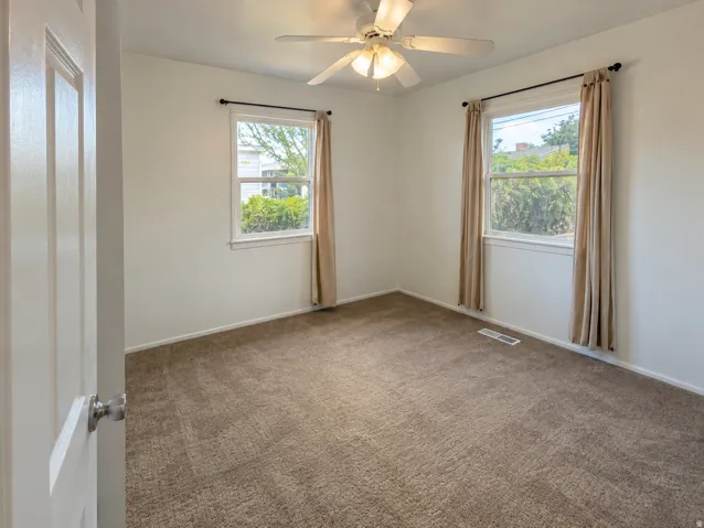 Neutral wall finishes, textured carpet flooring, two windows with trim, and a ceiling fan with integrated lighting