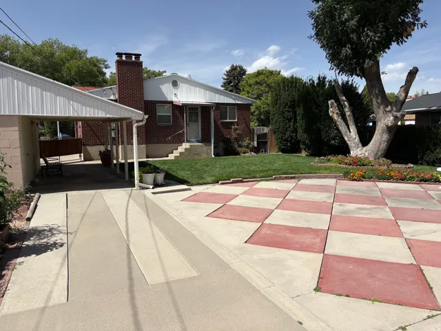 Brick exterior with a prominent chimney, covered carport, concrete driveway, and a checkered patio