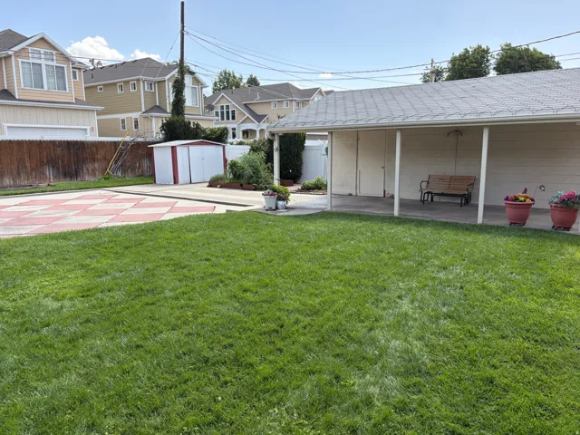 Expansive green lawn, covered patio area, concrete patio with patterned pavers, white storage shed, and wood fencing