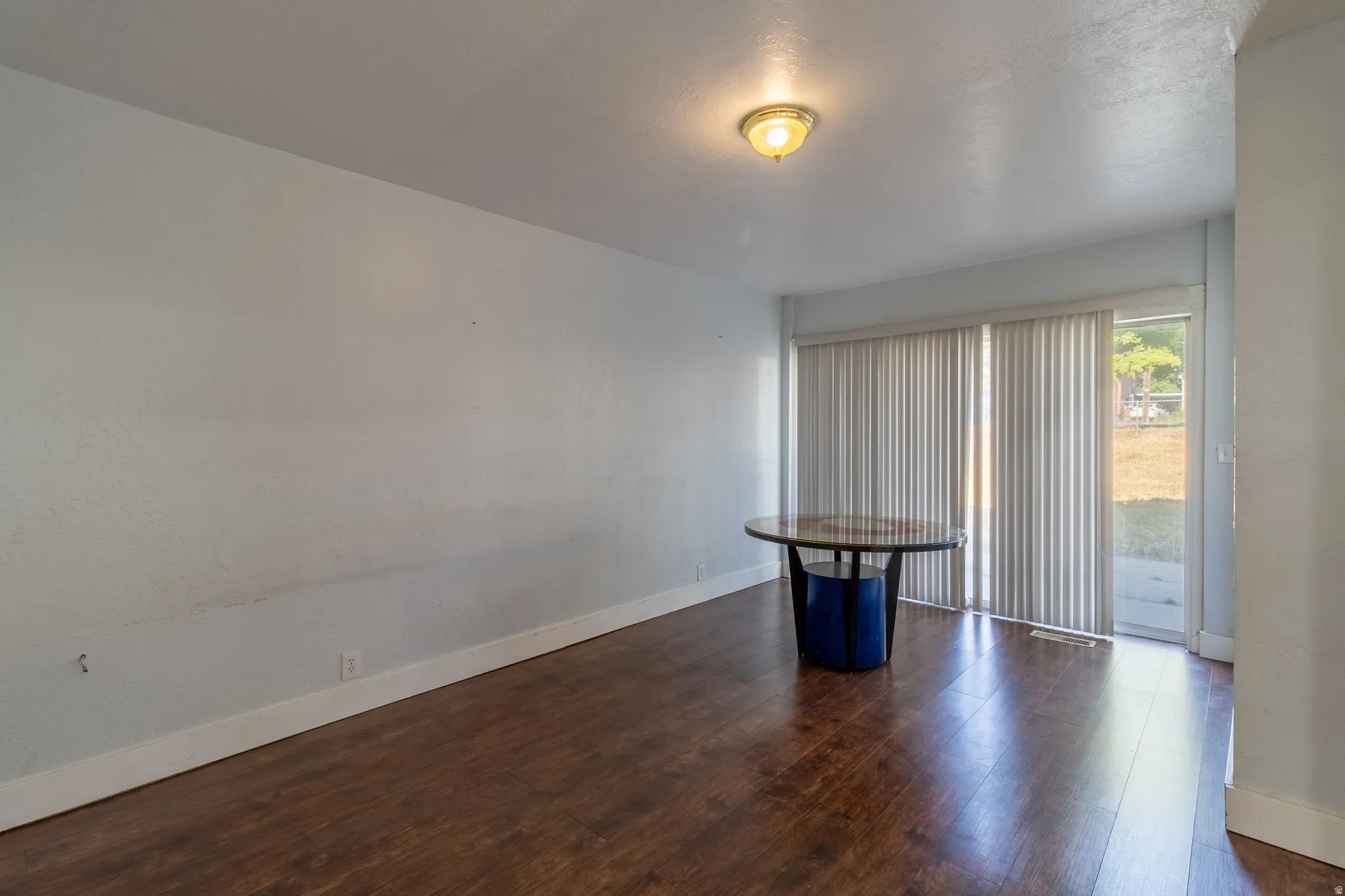 Wood-finish flooring, light-colored walls, ceiling-mounted light fixture, and vertical blinds on a sliding glass door