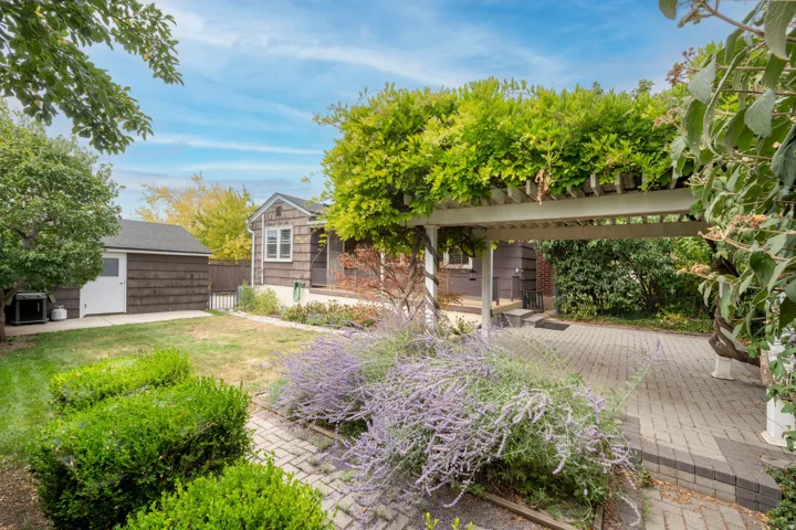 Paved patio with an overhead pergola, mature landscaping, and a detached shed