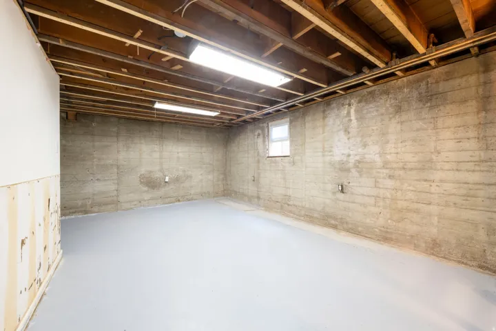 Exposed wood ceiling joists with overhead lighting, concrete foundation walls, painted concrete floor, and a single window