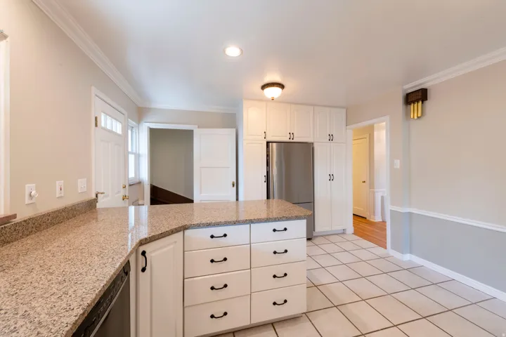 Kitchen featuring white cabinetry, light-toned granite countertops, stainless steel refrigerator, white tile flooring, and a central island with drawer storage