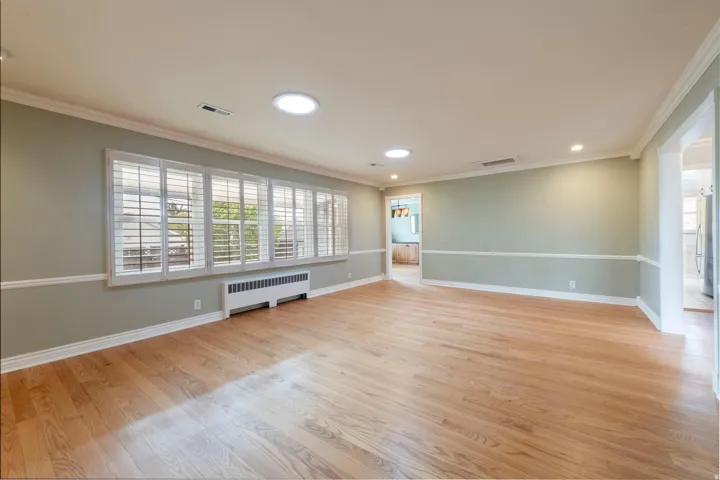 Spacious room featuring wood-finish flooring, white crown molding, recessed lighting, and a large window with white plantation shutters