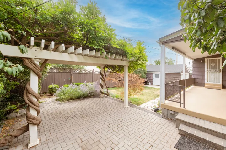 Paved patio area, white pergola structure, mature climbing vines, landscaped garden bed, and a covered porch with metal railing