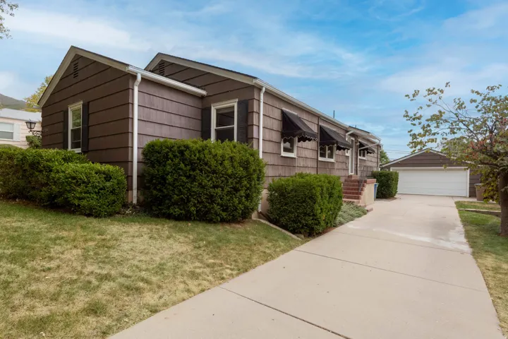 Brown horizontal siding exterior with black window shutters, three black awnings, a paved driveway, and a detached two-car garage with white door