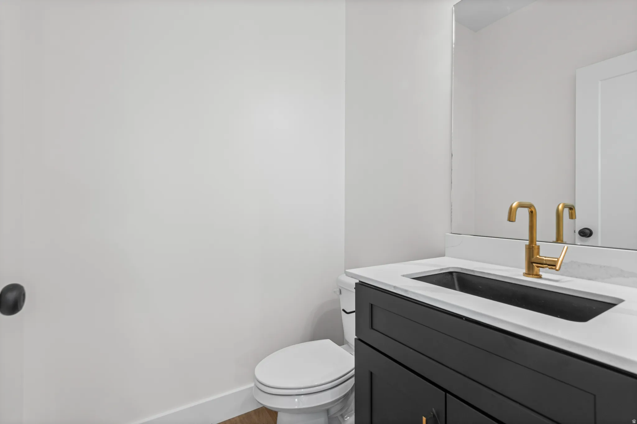 White countertop with an integrated dark sink, gold-finish faucet, dark vanity cabinetry, and a framed mirror