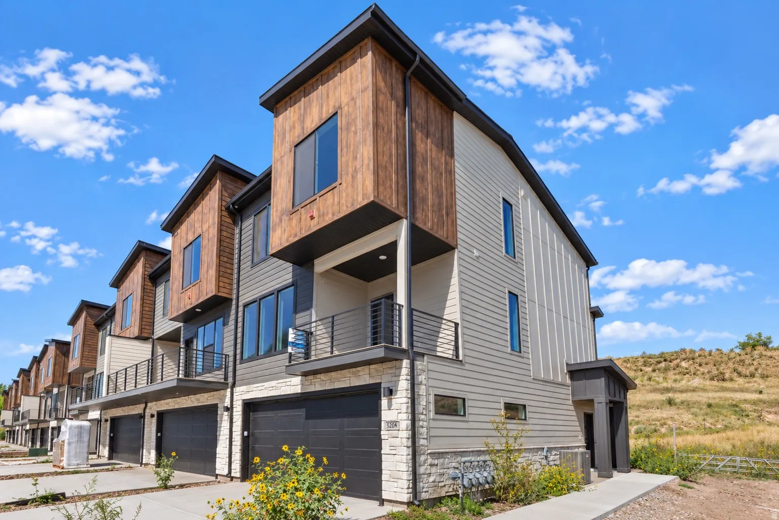 Modern architectural design featuring vertical wood-look siding, stone accents, multiple large windows, a private balcony with horizontal railings, and an attached two-car garage