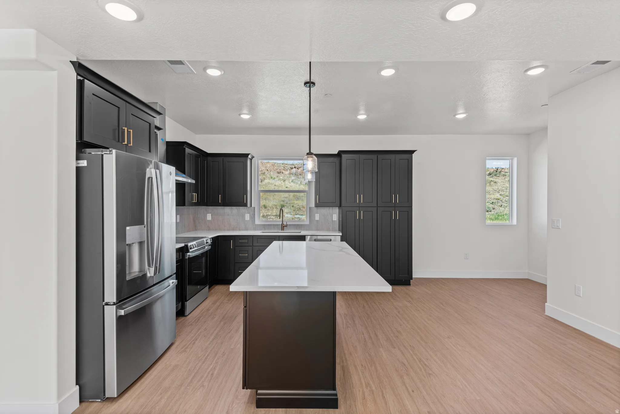 Kitchen featuring dark cabinetry, stainless steel appliances, a center island with a light-toned countertop, wood-finish flooring, and recessed lighting