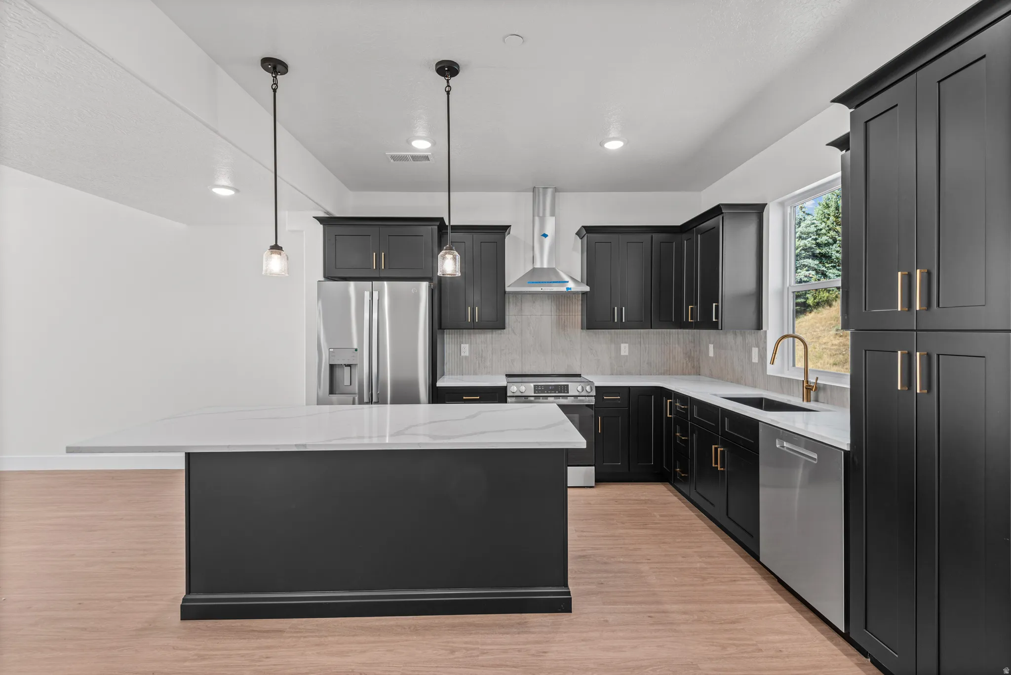 Kitchen featuring an island with a light-toned countertop, dark cabinetry with gold-toned hardware, stainless steel appliances, and wood-finish flooring