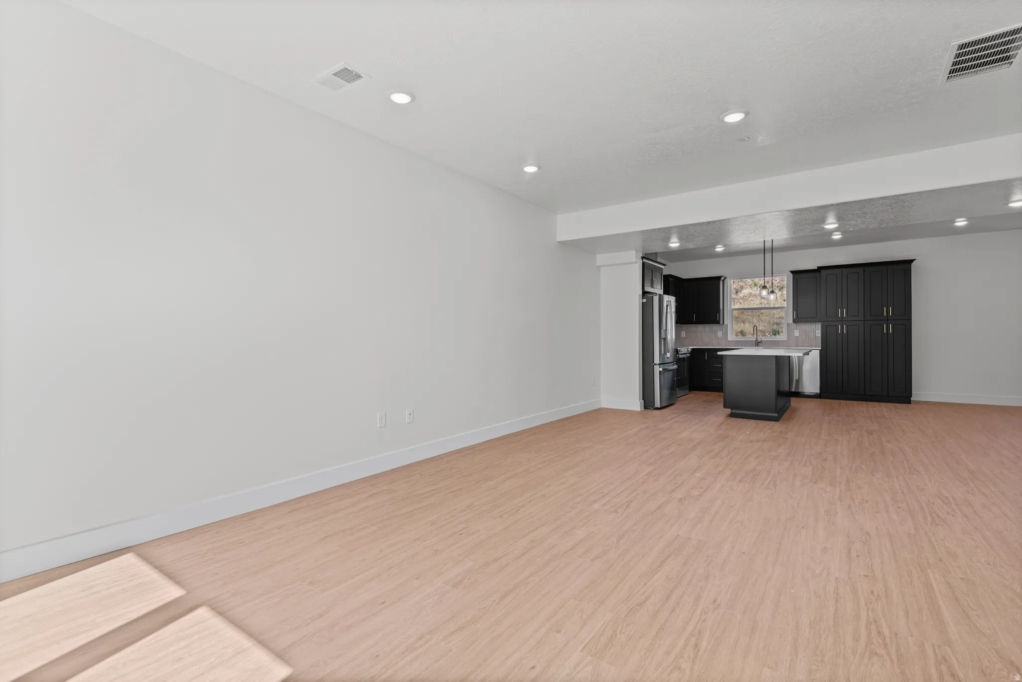Recessed lighting illuminates the open-concept space featuring wood-finish flooring, a kitchen island, dark cabinetry, and stainless steel appliances