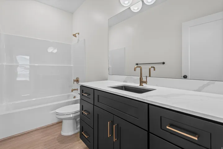 Wood-finish flooring, white tub and shower surround, black vanity with white countertop, gold-toned faucet, and rectangular undermount sink