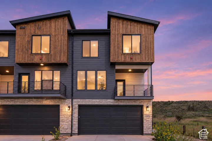 Modern multi-level exterior featuring wood-tone paneling, dark horizontal siding, stone veneer masonry, dual balconies with metal railings, and attached multi-car garages