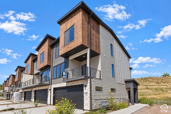 Modern architectural design featuring vertical wood-look siding, stone accents, multiple large windows, a private balcony with horizontal railings, and an attached two-car garage