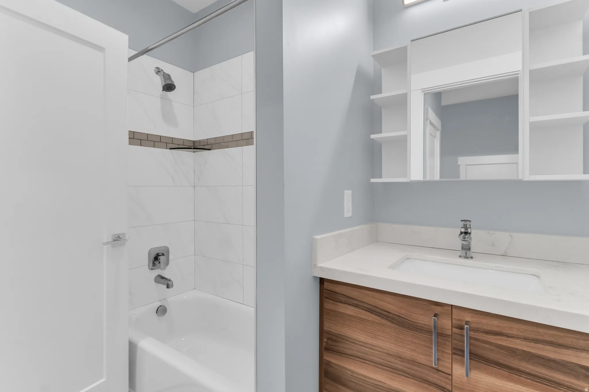 Bathroom featuring a white bathtub with a tiled shower surround, a wood-finish vanity with an integrated sink, a light-colored countertop, and a mirrored medicine cabinet with open shelving