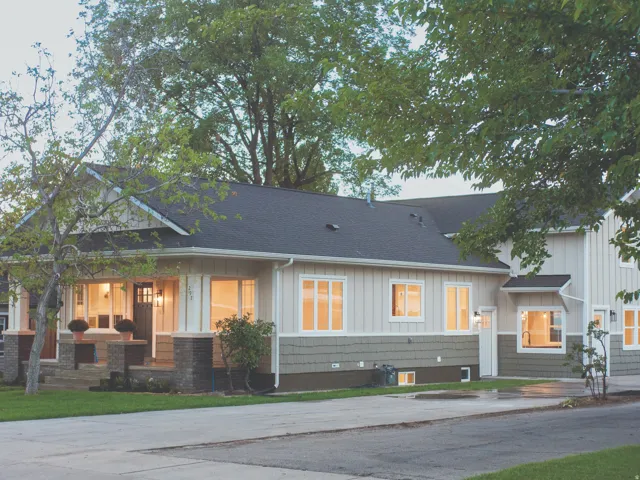 Modern bungalow exterior showcasing a light gray and white board-and-batten facade with dark asphalt shingle roofing, expansive front-facing windows with warm interior lighting, a covered front porch with brick foundation detailing, and mature landscaping