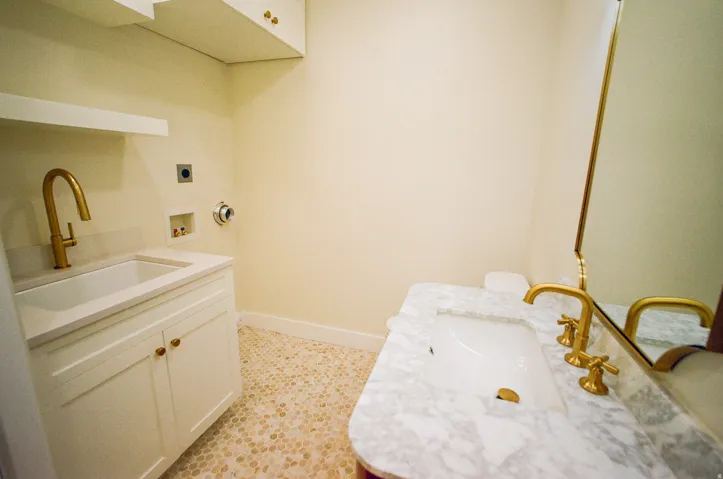 Integrated laundry sink with a brass-finish faucet, white cabinetry with brass-finish hardware, upper built-in cabinetry, and a marble-finish countertop vanity with a brass-finish faucet