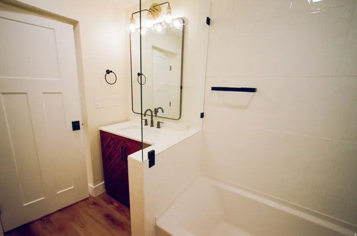 Wood-finish flooring, white paneled door, custom vanity with integrated sink, gold-toned wall-mounted light fixture, and tiled shower surround with glass partition