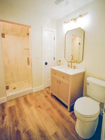 Wood-finish flooring, glass-enclosed shower with pebble-finish base, vanity with integrated sink, gold-tone fixtures, and an illuminated mirror