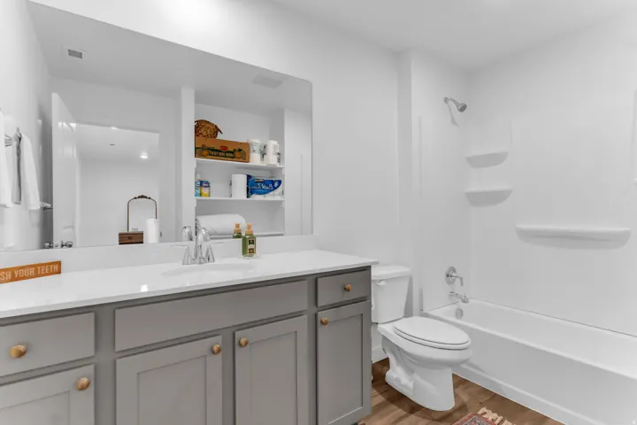 Bathroom featuring a vanity with a white countertop and undermount sink, gray cabinetry with brass-toned hardware, a full-width mirror, and a white tub/shower combination with built-in shelving