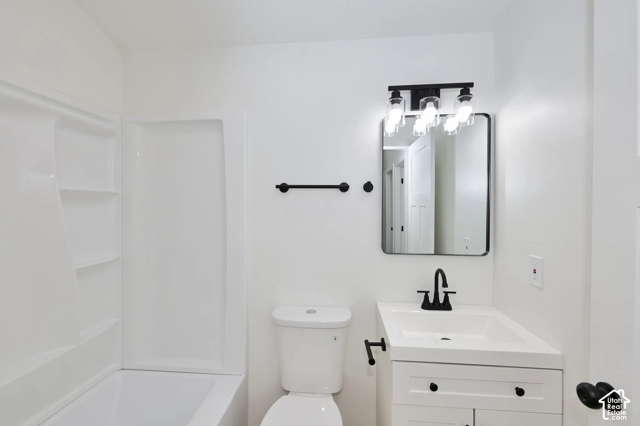 White bathtub with integrated shelving, matte black vanity light fixture, rectangular mirror with a black frame, white vanity with black hardware, and a black faucet