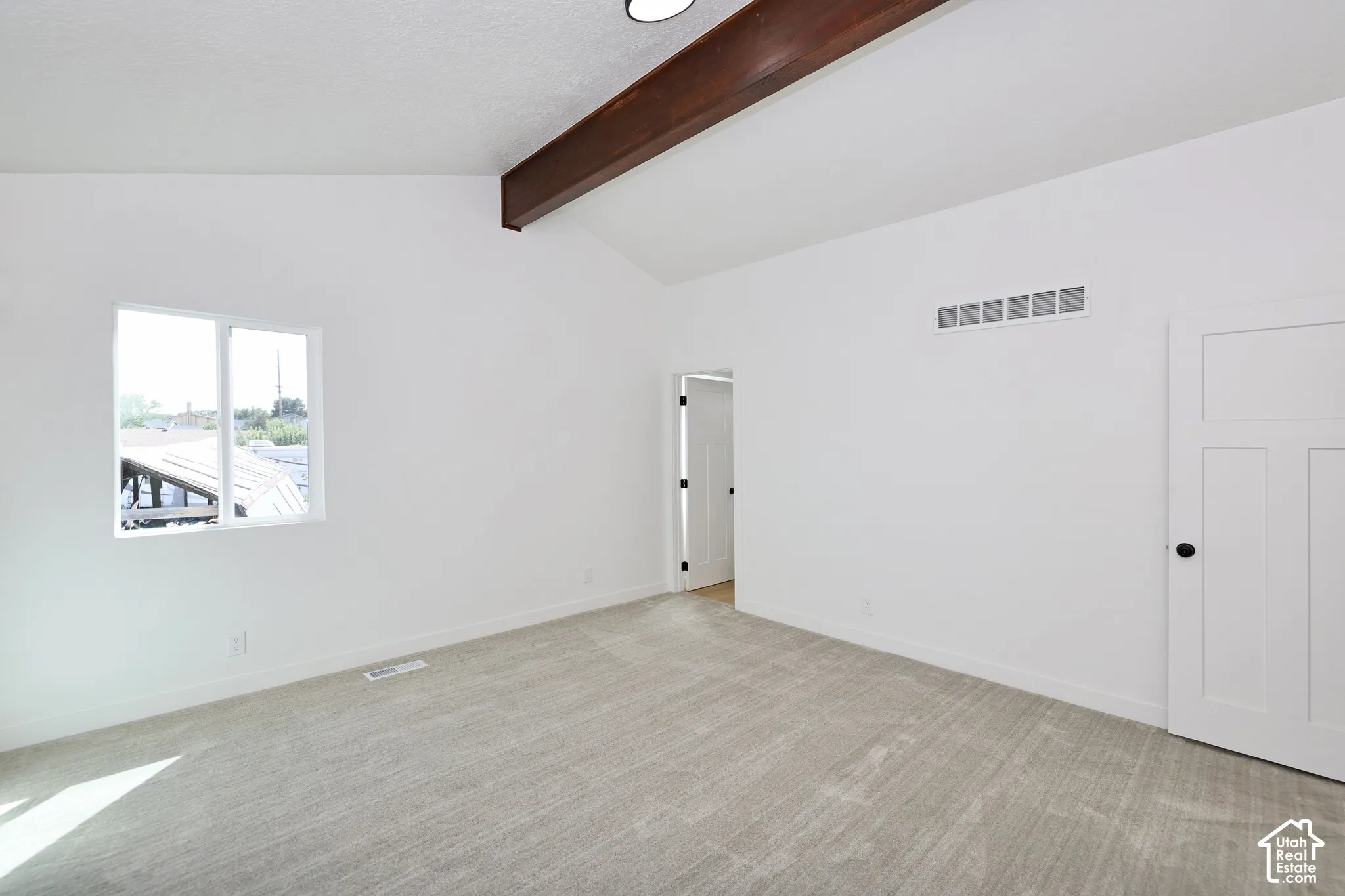Vaulted ceiling featuring an exposed beam, light gray carpet, white interior door with black hardware, and a single window