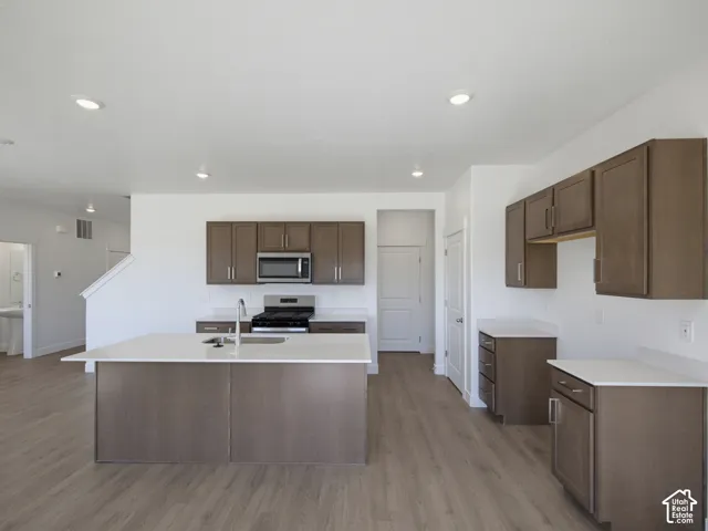 Kitchen featuring a central island with an integrated sink, wood-finish flooring, white countertops, dark wood-finish cabinetry, and stainless steel appliances