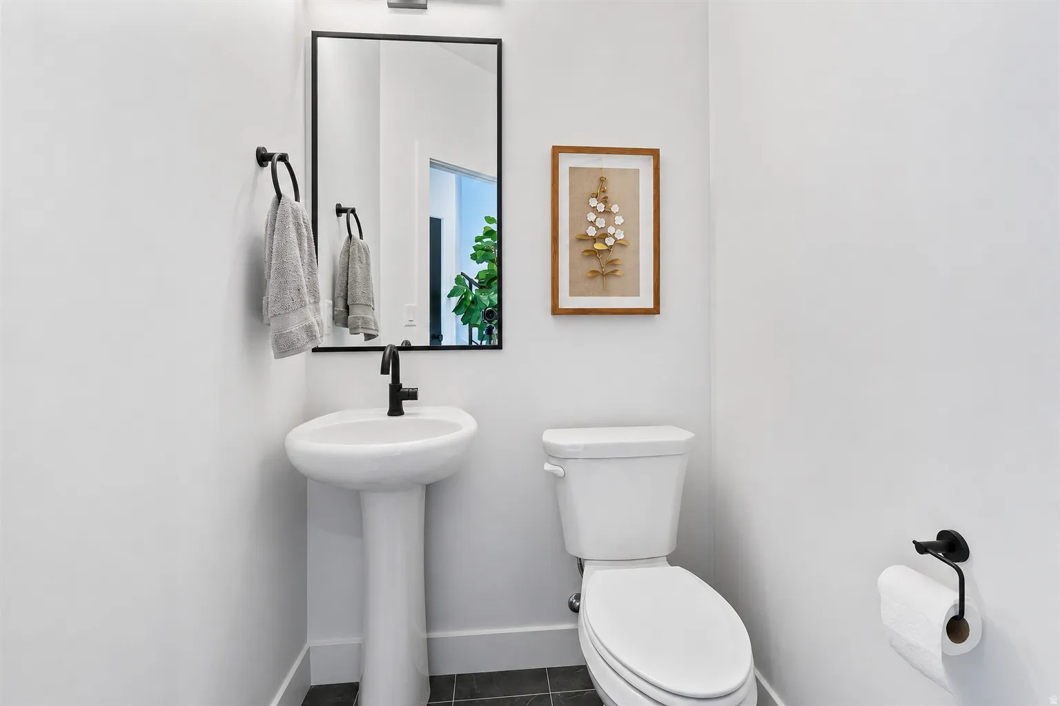 Pedestal sink with a matte black faucet, rectangular mirror with a black frame, and dark tile flooring