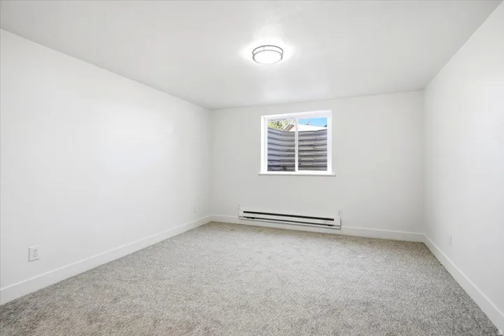Carpeted flooring, white painted walls, ceiling-mounted light fixture, window with white trim, and baseboard heater