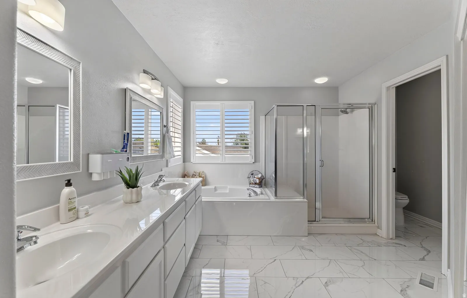 Dual vanity with integrated sinks, framed mirrors, glass-enclosed shower, soaking tub, and white plantation shutters
