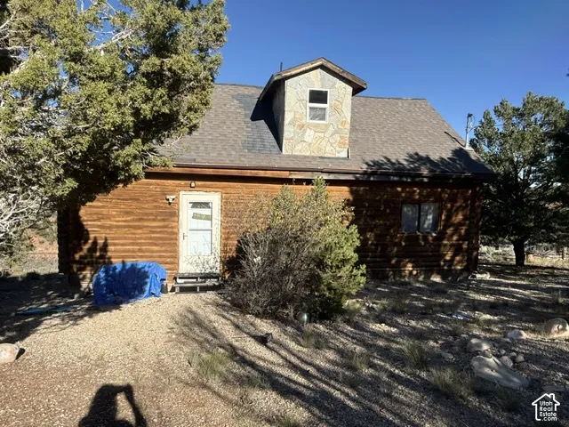 Log exterior cabin featuring a stone-clad dormer, shingled gable roof, and natural gravel grounds
