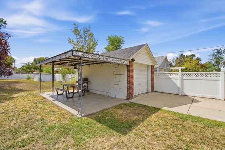 Detached garage with white overhead door and brick accents, concrete driveway, white lattice-top fencing, and an ornate metal pergola over a concrete patio
