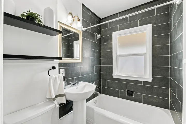 Pedestal sink, bathtub with shower, dark subway tile surround, and a framed mirror