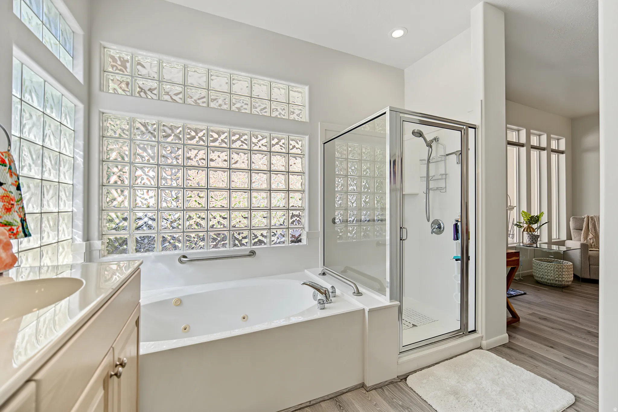 Built-in jetted tub with grab bars, glass block windows, separate glass-enclosed shower, wood-finish flooring, and recessed lighting