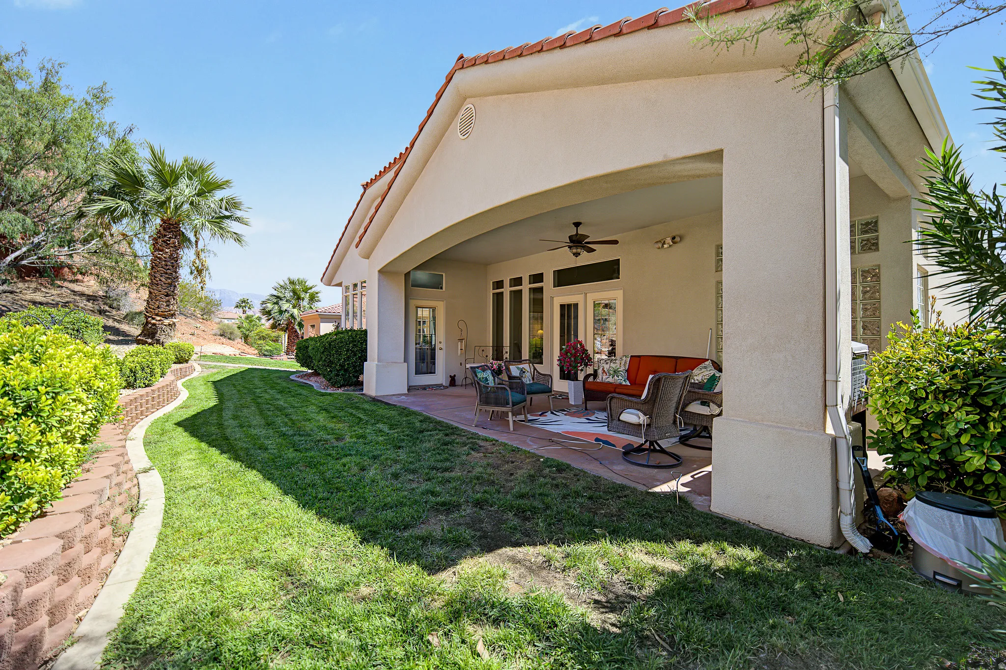 Covered patio with a ceiling fan and exterior lighting, featuring multiple French doors, an arched stucco facade, and a terraced retaining wall