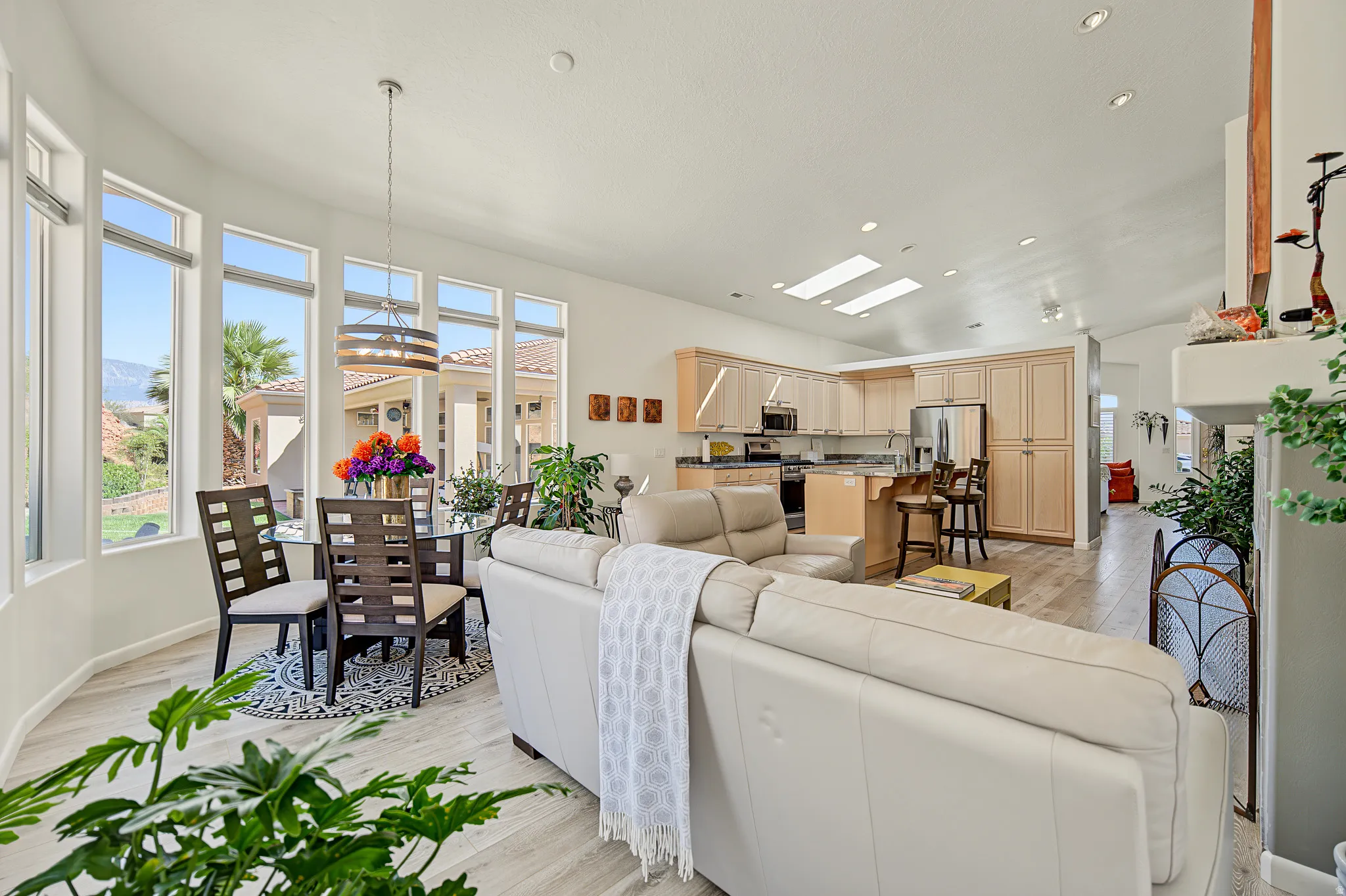 Open-concept living space featuring wood-finish flooring, a kitchen with light wood cabinetry, recessed lighting, and skylights, and a dining area with a contemporary chandelier and multiple windows