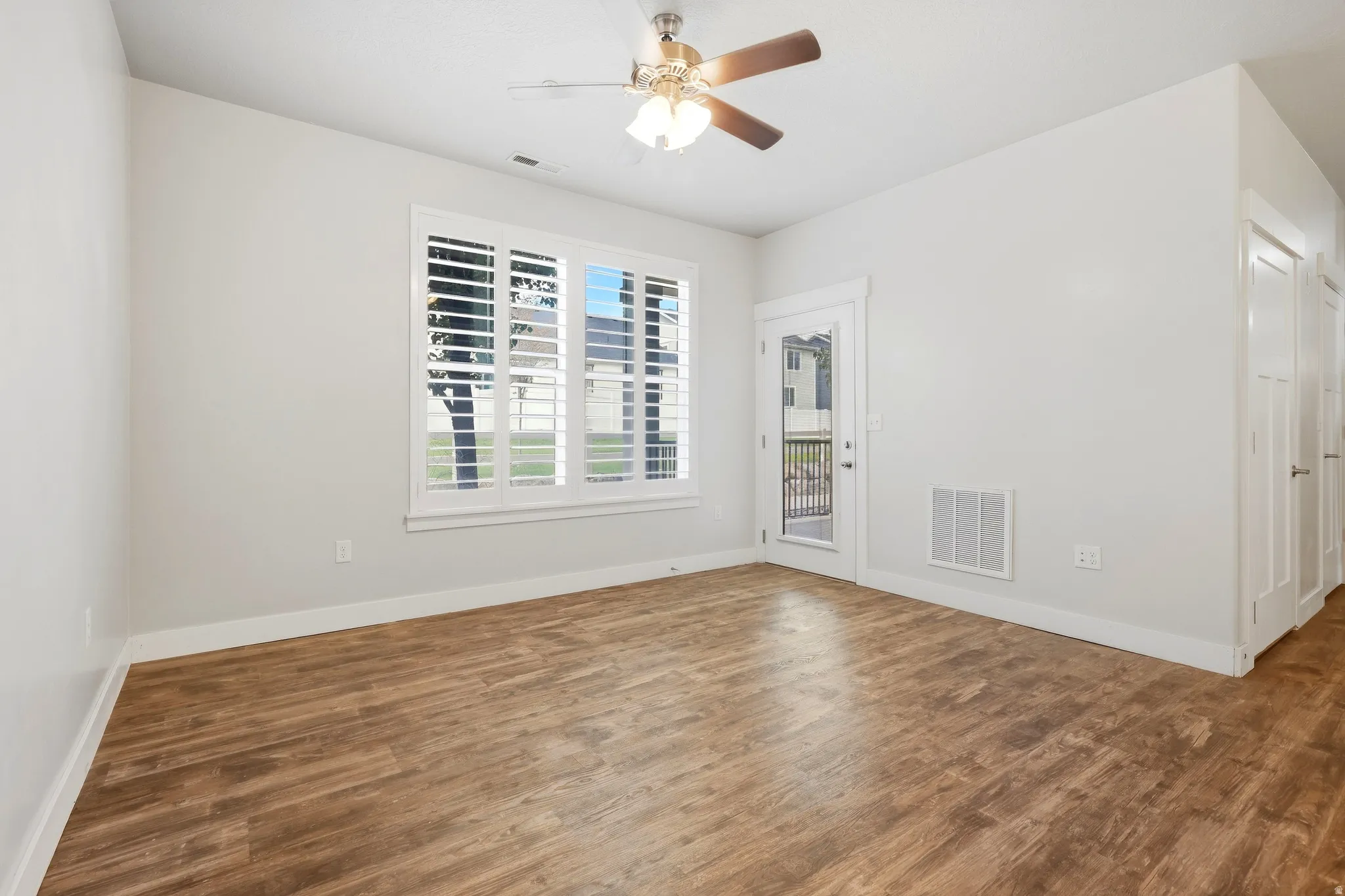 Wood-finish flooring, ceiling fan with integrated lighting, white plantation shutters, glass-paneled exterior door, and white interior doors