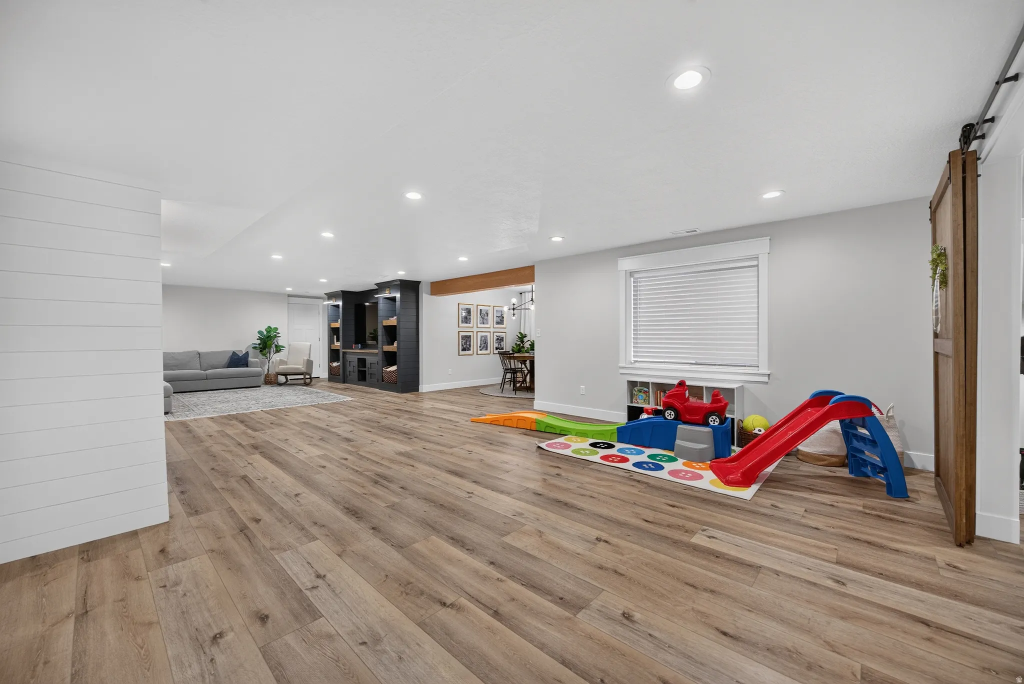 Expansive open-concept room featuring wood-finish flooring, recessed lighting, a built-in media center, a window with blinds, and shiplap wall accents