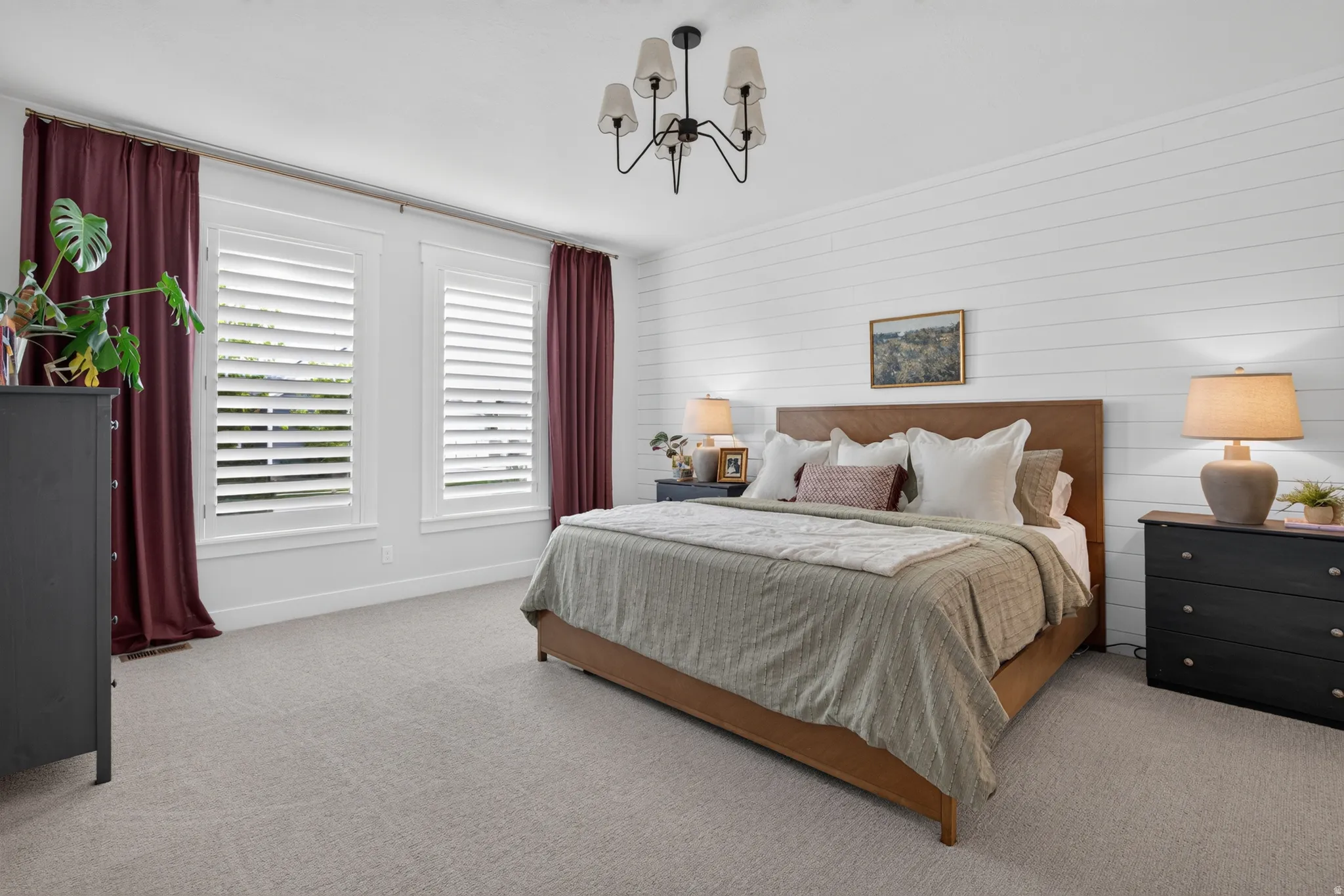 White shiplap accent wall, two large windows with white plantation shutters, light gray carpeting, and a ceiling-mounted chandelier