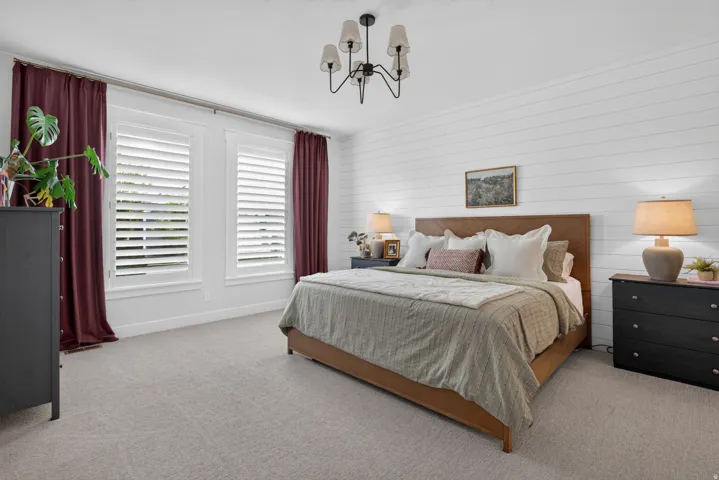 White shiplap accent wall, two large windows with white plantation shutters, light gray carpeting, and a ceiling-mounted chandelier