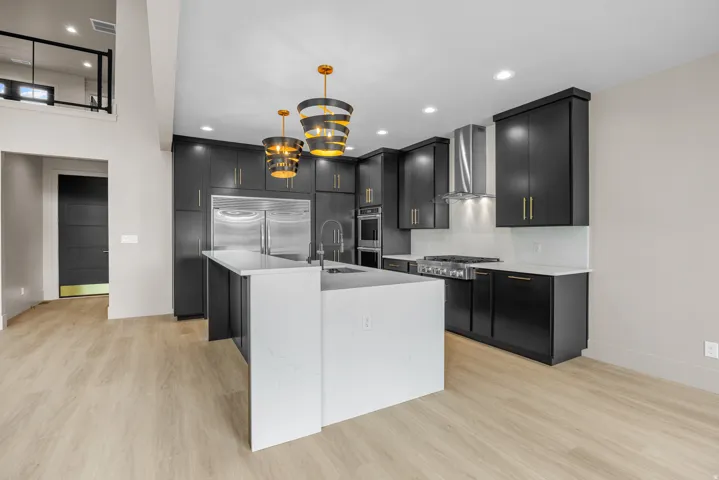 Modern kitchen featuring an expansive island with a waterfall edge, dark cabinetry with brass hardware, stainless steel appliances, and wood-finish flooring