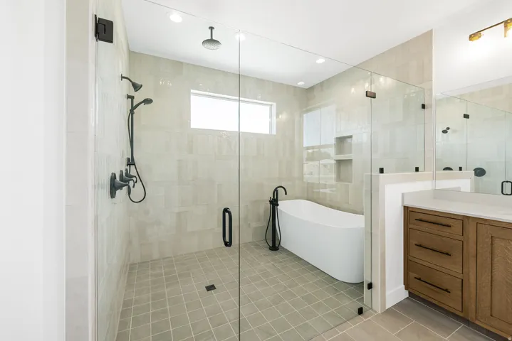 Glass-enclosed shower with multiple showerheads, freestanding soaking tub with floor-mounted faucet, wood-finish vanity with countertop, and recessed lighting