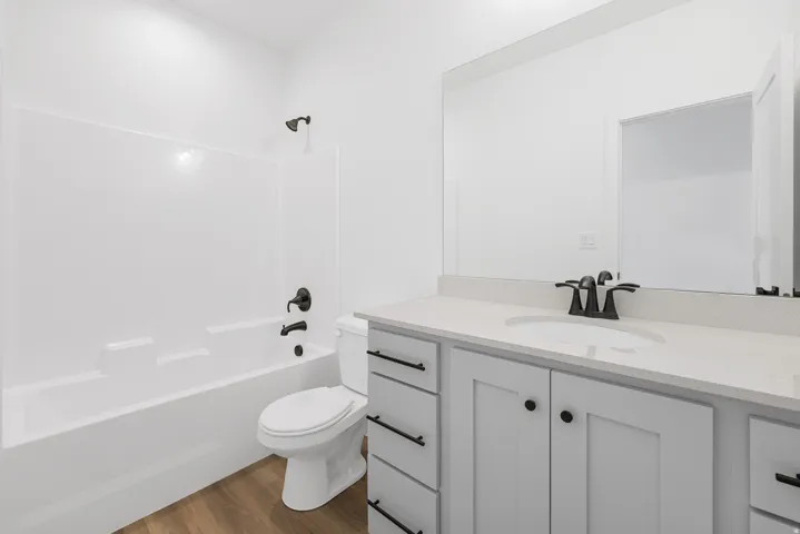White integrated tub and shower unit, wood-finish flooring, white vanity with stone-finish countertop, single basin sink, and matte black hardware