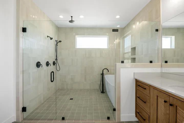 Spacious bathroom featuring a frameless glass shower enclosure, matte black fixtures, a freestanding soaking tub, and a wood-finish vanity with a light-toned countertop