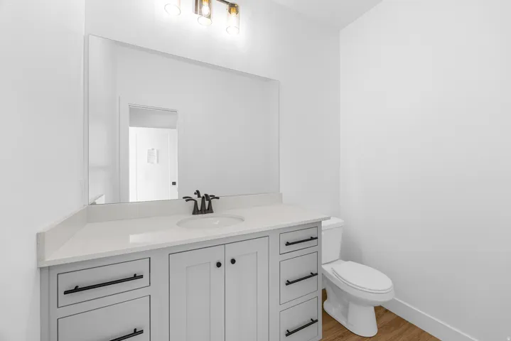 White vanity with black hardware, white countertop with integrated sink, rectangular mirror, three-light vanity fixture, and wood-finish flooring