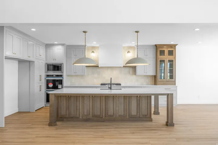 Kitchen featuring light wood-finish flooring, two-tone cabinetry, a built-in oven and microwave, a center island with a sink, and pendant lighting