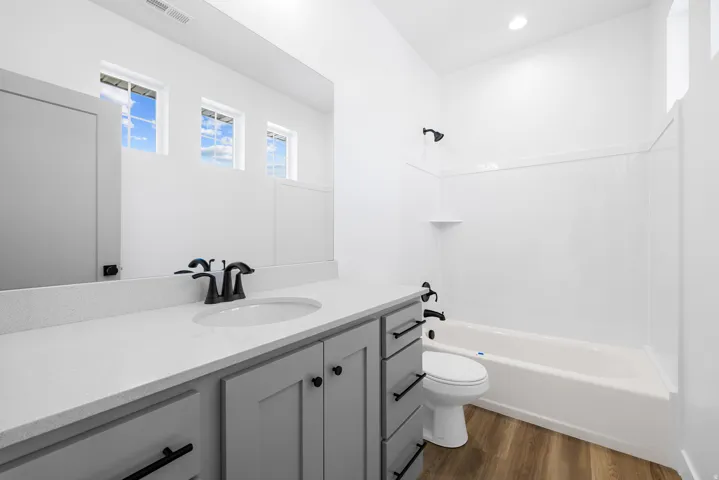 Wood-finish flooring, white bathtub with shower, light gray vanity with black hardware, white countertop, and three transom windows