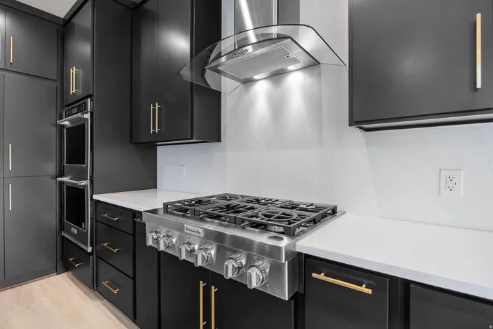 Matte black cabinetry with gold-tone hardware, stainless steel gas range, double wall ovens, white countertop, and range hood with integrated lighting