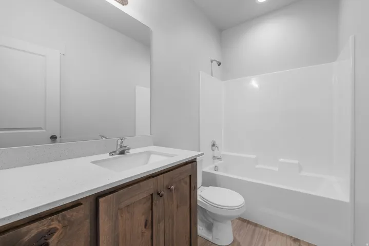 Vanity with wood-finish cabinetry and light-toned countertop, integrated sink with chrome faucet, full wall mirror, white bathtub with shower, and wood-finish flooring